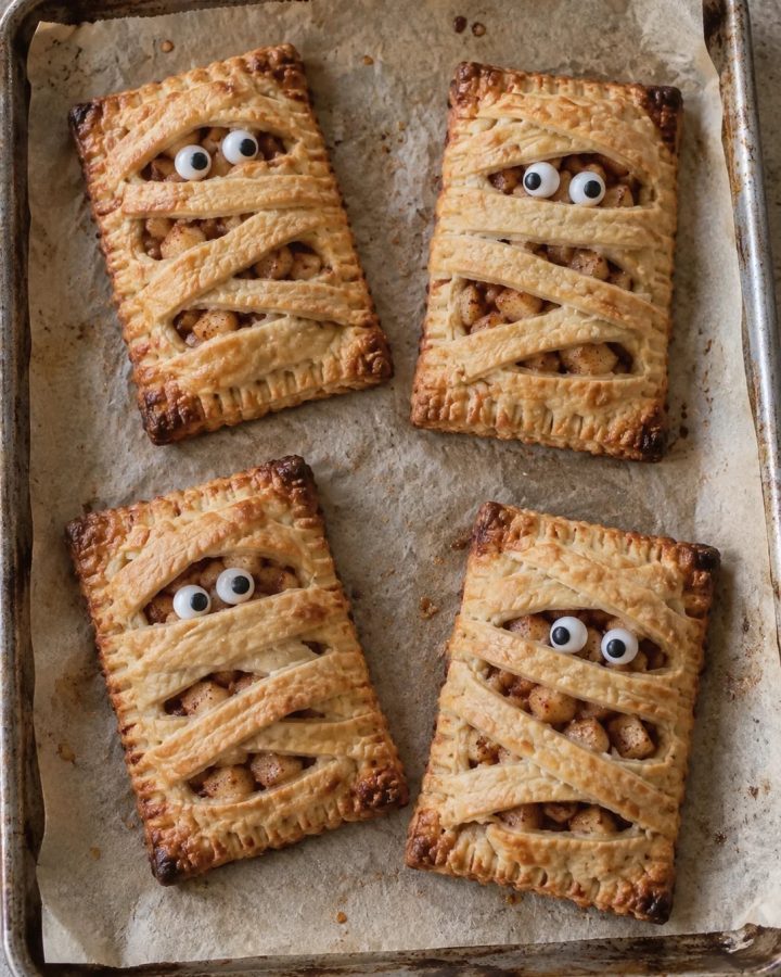 Generated illustration of cinnamon-apple mummy hand pies with crisscrossed golden pastry strips and candy eyes on parchment.